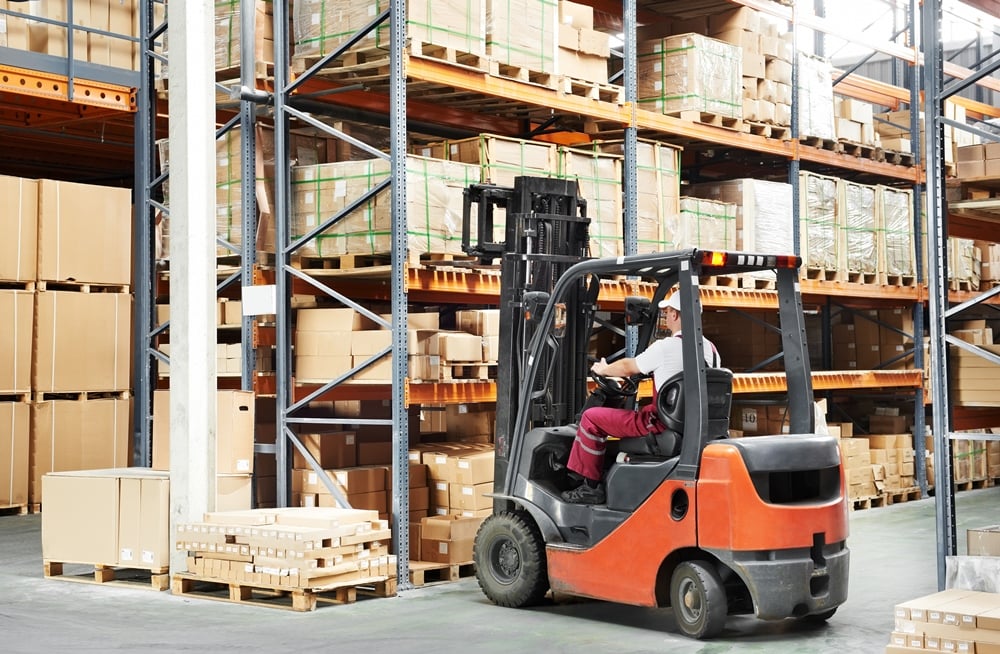 Forklift operator navigating through a warehouse with stacked cardboard boxes and shelving units.
