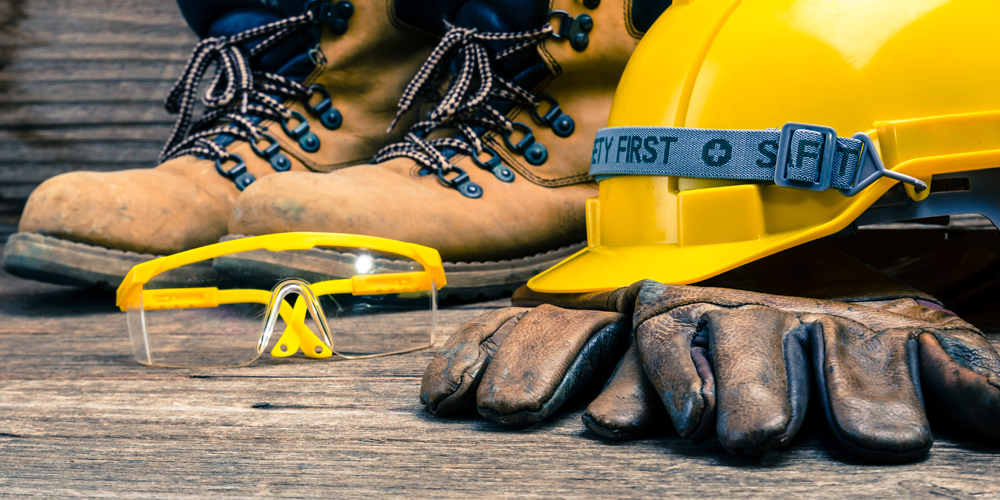 Safety gear including work boots, gloves, safety goggles, and a hard hat situated on a wooden surface.