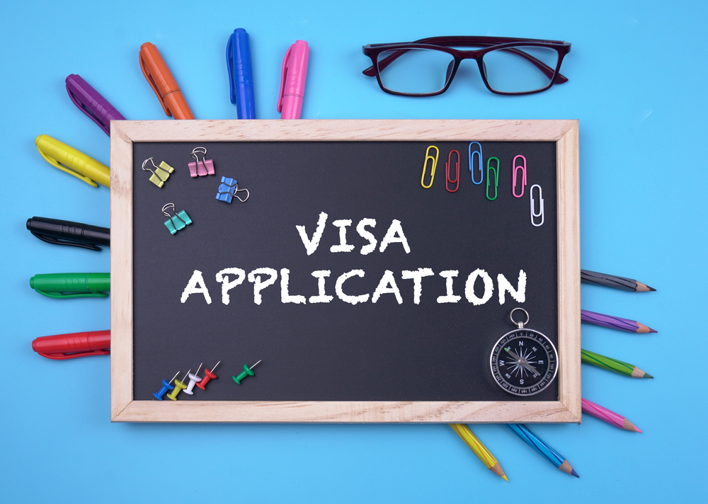 Chalkboard displaying "VISA APPLICATION" surrounded by colorful pens, paperclips, and a compass.