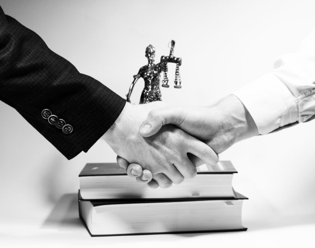 Hands of two individuals shaking, with law books and a scales of justice statue in the background.