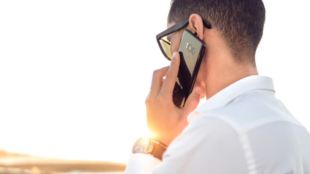 Person in sunglasses holding a smartphone to their ear while standing outdoors in sunlight.