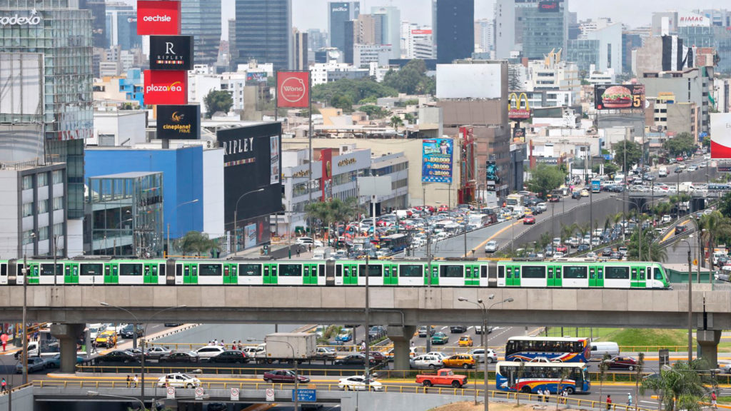 Urban landscape featuring a green and white elevated train passing over busy streets filled with vehicles and commercial buildings.