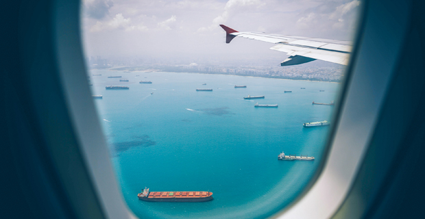 Aerial view of cargo ships anchored in blue waters beneath an airplane wing.