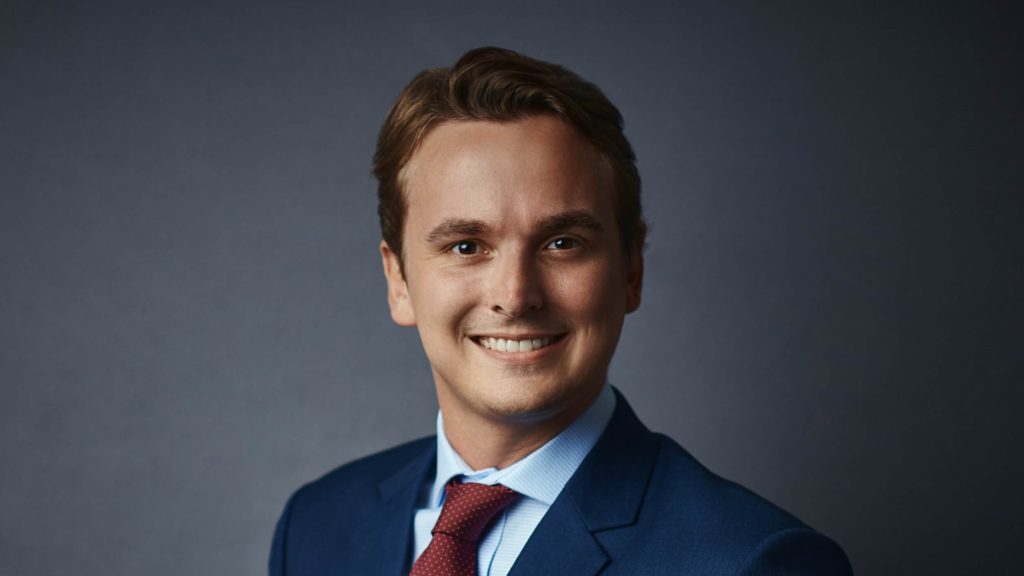 Young professional in a suit smiling against a neutral background, representing a lawyer or legal consultant.