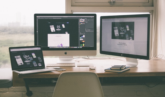 Desk with a desktop computer and laptop displaying website designs, alongside a notepad and smartphone.