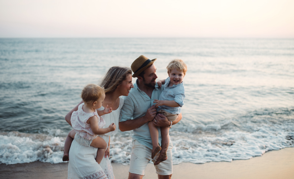Family enjoying a playful moment together at the beach, with children being carried by their parents near the water.
