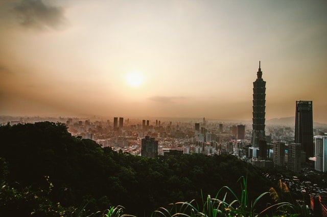 City skyline at sunset with modern skyscrapers, including Taipei 101, against a hazy, colorful sky.