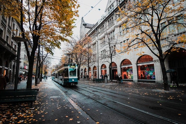 Tram driving along a wet city street lined with trees and historic buildings, with pedestrians walking nearby.