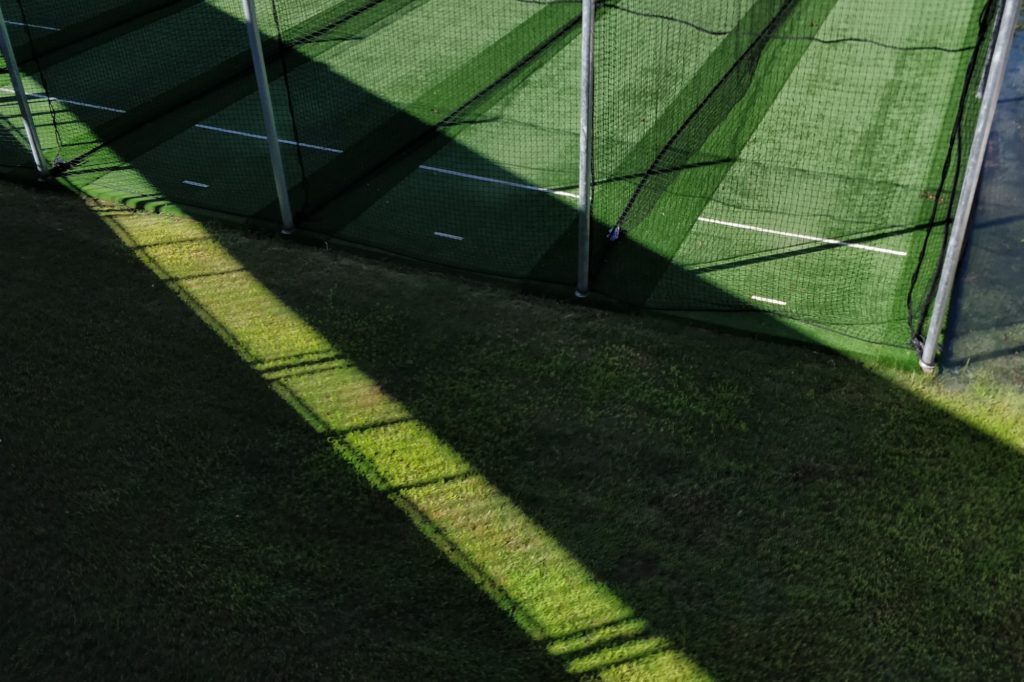 Green turf field with long shadows cast by netting and fencing.