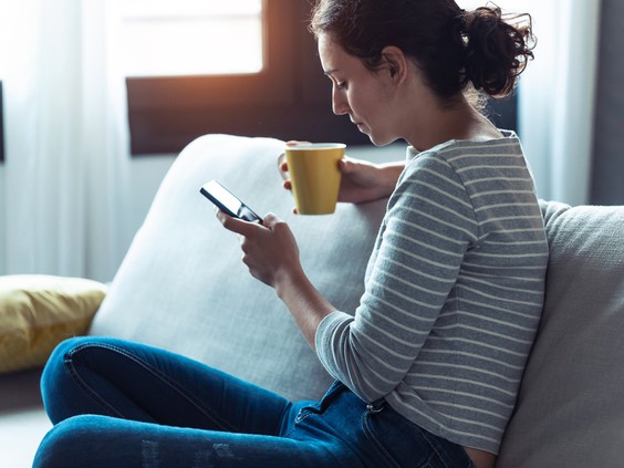 Woman sitting on a couch, holding a coffee cup while looking at a smartphone in a bright, cozy living room.