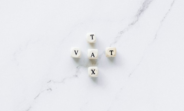 White dice with letters spelling "VAT" and "X" arranged on a marble surface.