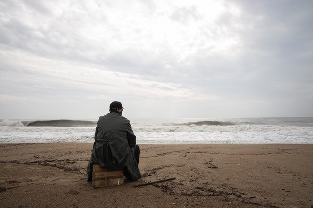 Person sitting on a wooden crate on a beach, looking out at ocean waves under a cloudy sky.