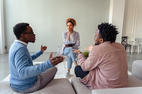 Mediator facilitating discussion between two men in a neutral setting, seated on a sofa with notepad in hand.