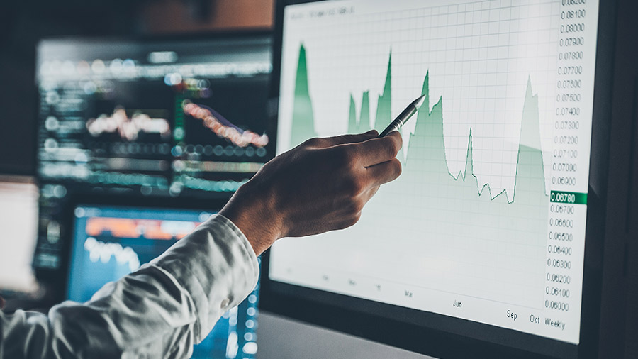 Hand pointing at data trends on financial graph displayed on computer screen in an office setting.