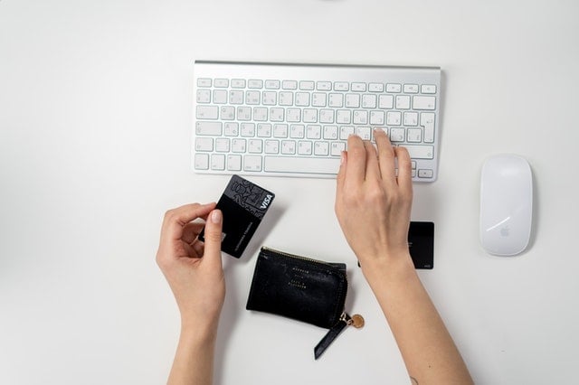 Hand holding a credit card above a keyboard, with a wallet and another card visible on a white desk.