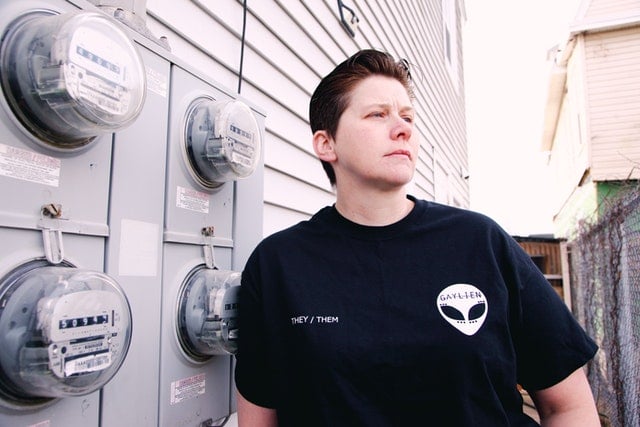 Individual standing next to electrical meters, wearing a black shirt with "THEY/THEM" text.