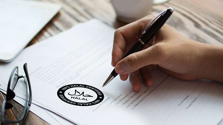 Hand holding a pen reviewing documents with a '100% HALAL CERTIFIED' sticker on the table.