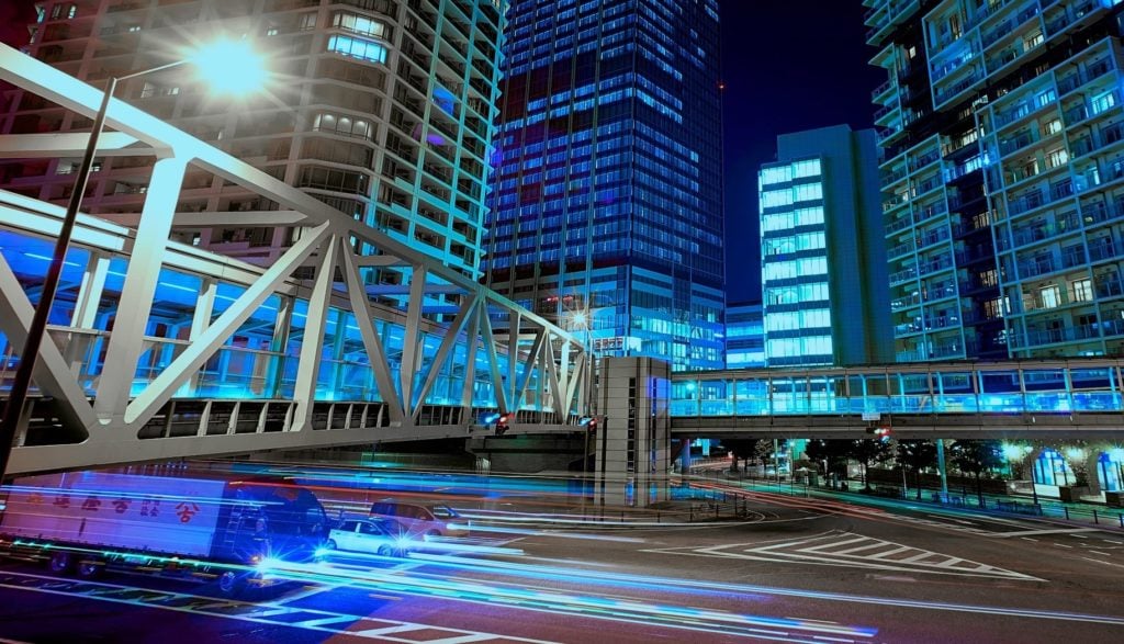 Urban night scene with traffic and illuminated modern buildings, featuring pedestrian bridge and busy road.