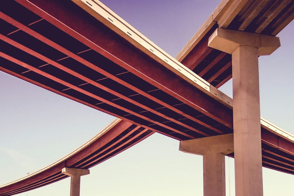 Overhead view of intersecting highway overpasses supported by concrete pillars against a clear sky.