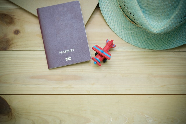 Brown passport next to a toy airplane and a woven hat on a wooden surface.