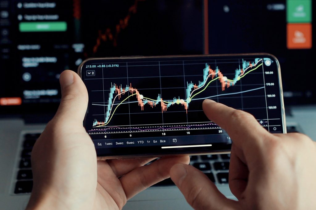 Person analyzing financial data on smartphone with stock market graphs displayed, laptop screen in background.