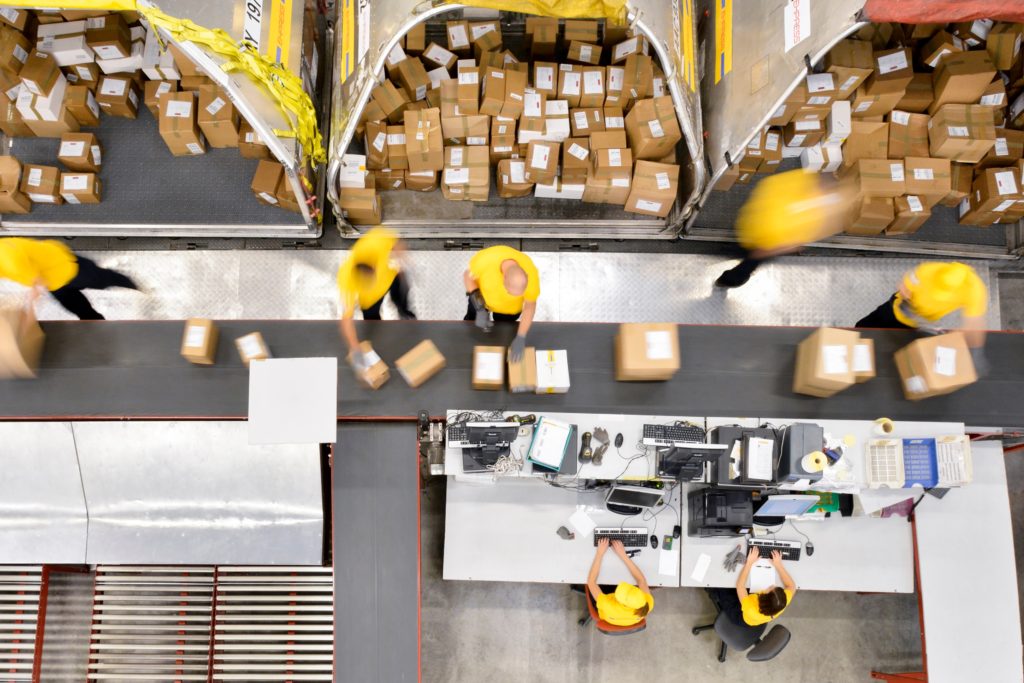 Workers sorting packages on a conveyor belt at a distribution center with shipping stations in view.