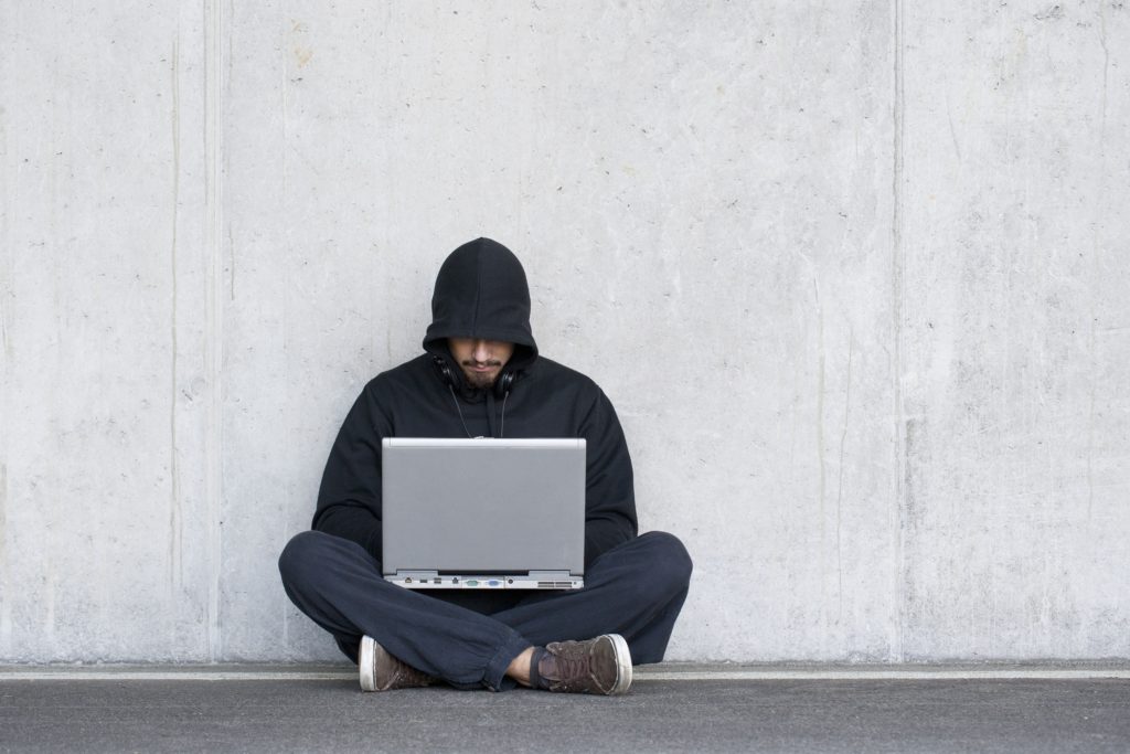 Individual sitting on the ground with a laptop, wearing a hoodie and headphones, in a neutral environment.