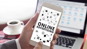 Person holding smartphone displaying "Online Advertising" with icons, in front of a laptop and coffee cup.
