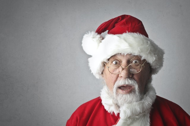 Surprised man in festive Santa costume with glasses and white beard against a plain gray background.