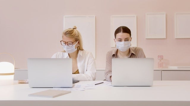 Two women wearing masks working on laptops at a desk with legal documents and stationery in a clean, modern workspace.