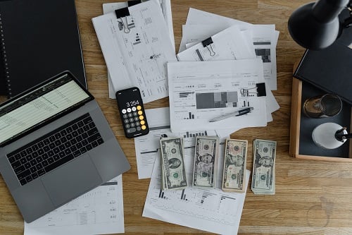 Laptop, calculator, financial documents, and cash arranged on a work desk in a professional setting.