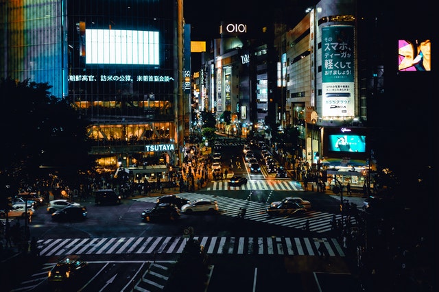 Busy urban intersection at night with vehicles and pedestrians, surrounded by bright signage and commercial buildings.
