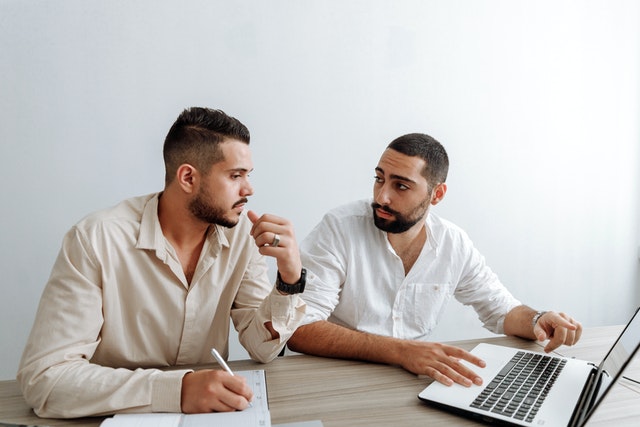 Two men discussing strategies while reviewing documents in front of a laptop at a desk.