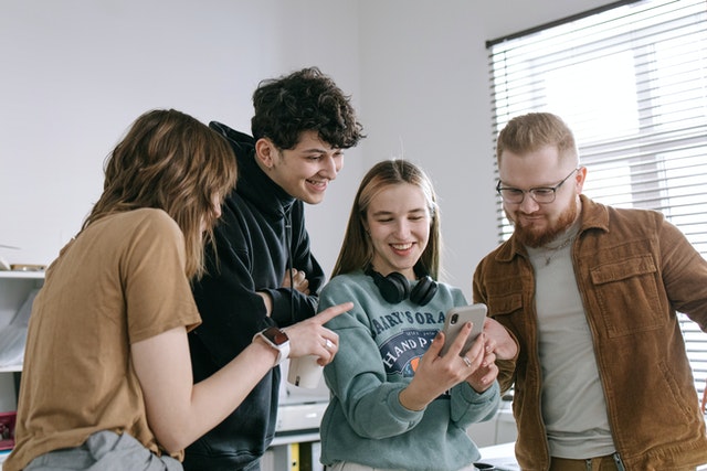 Group of four young adults gathered around a smartphone, smiling and engaging in conversation in a casual setting.