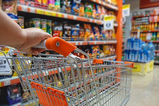 Person holding shopping cart in aisle with various grocery items displayed on shelves.
