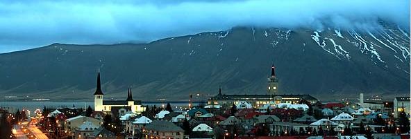 Cityscape with mountains in background, featuring church steeples and illuminated buildings at dusk.