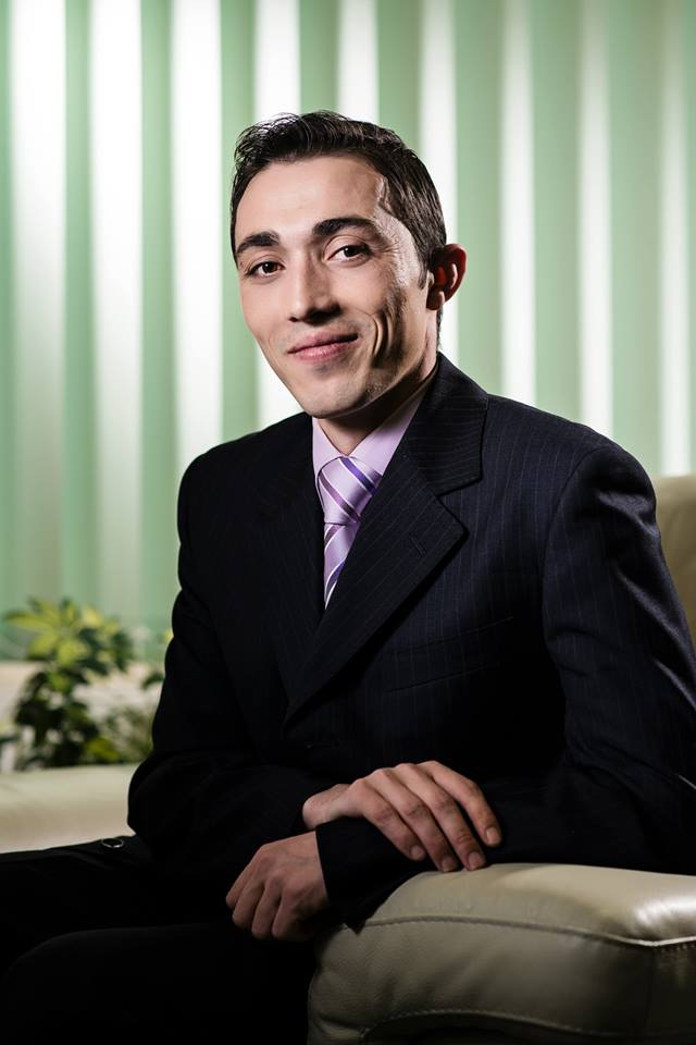 Lawyer seated in office, wearing a suit and tie, with a confident expression against a backdrop of green curtains.