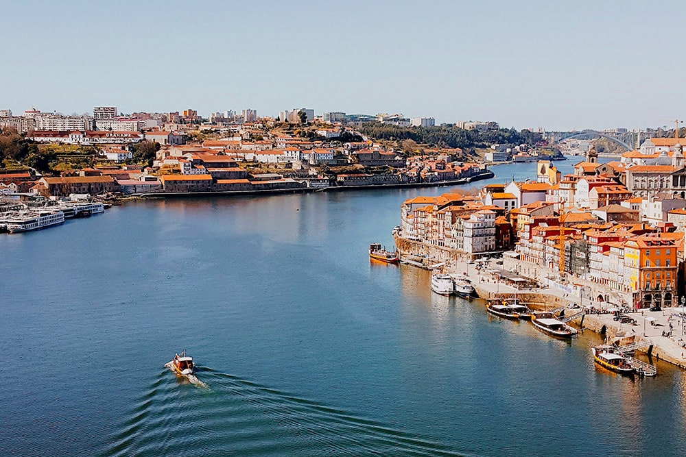 Cityscape of riverside buildings with orange rooftops and boats on a calm river under a clear blue sky.