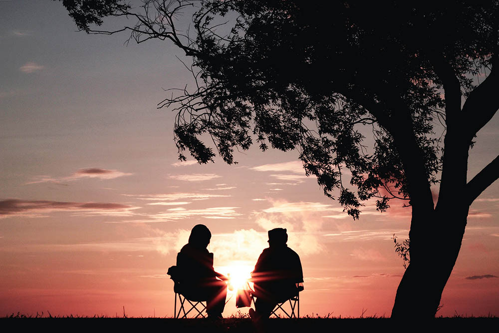 Couple sitting on chairs under a tree, watching a sunset with warm colors in the sky.