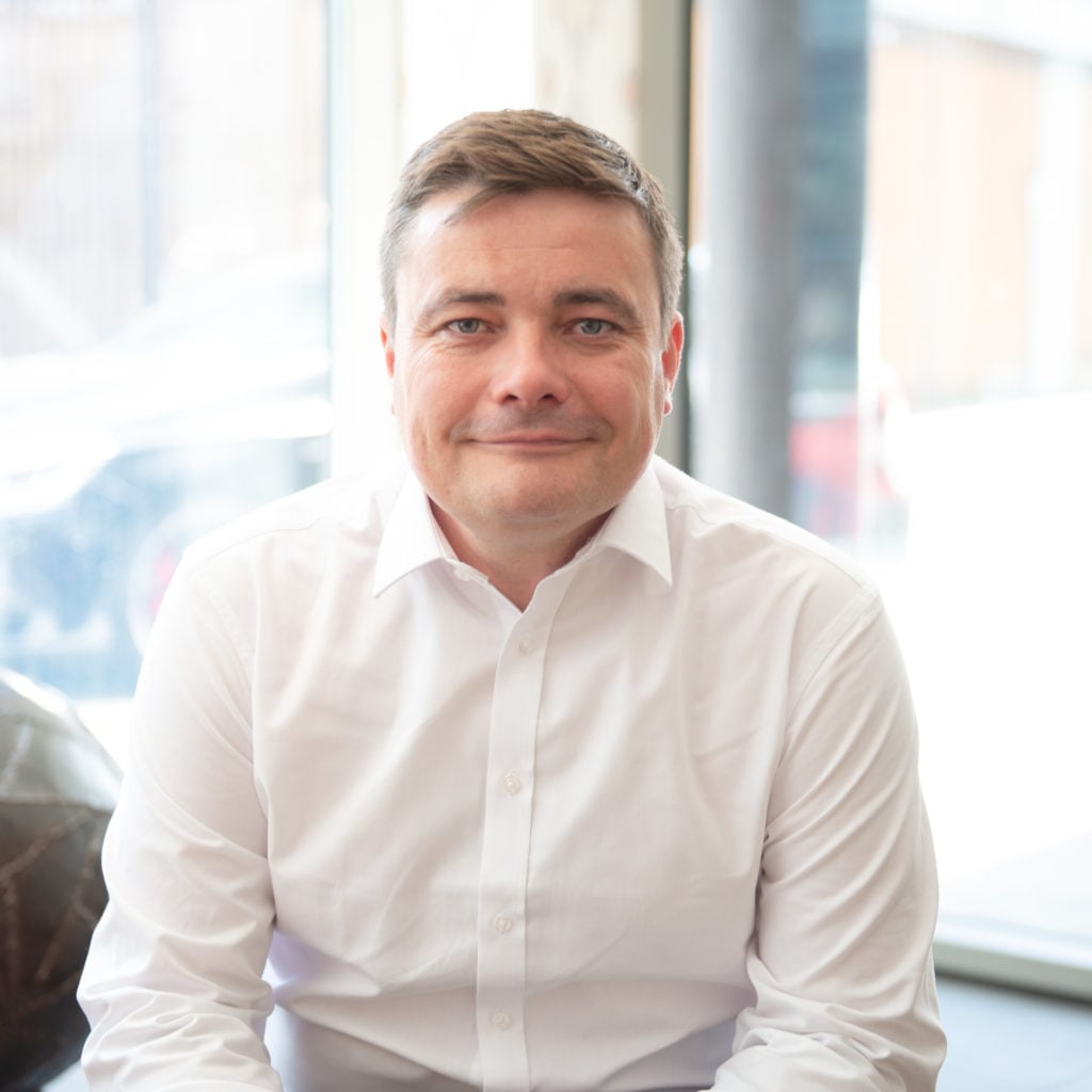 Legal professional seated indoors with a neutral expression, wearing a white shirt, in a modern office setting.