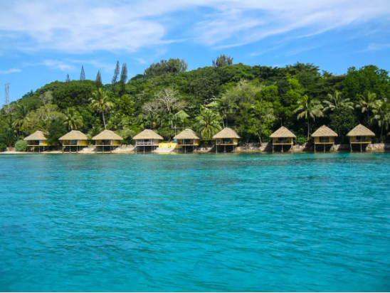 Tropical beachfront with thatched-roof bungalows along clear blue water and lush green vegetation in the background.