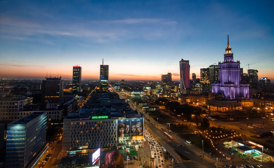City skyline at dusk featuring modern buildings, illuminated streets, and a prominent historical tower.