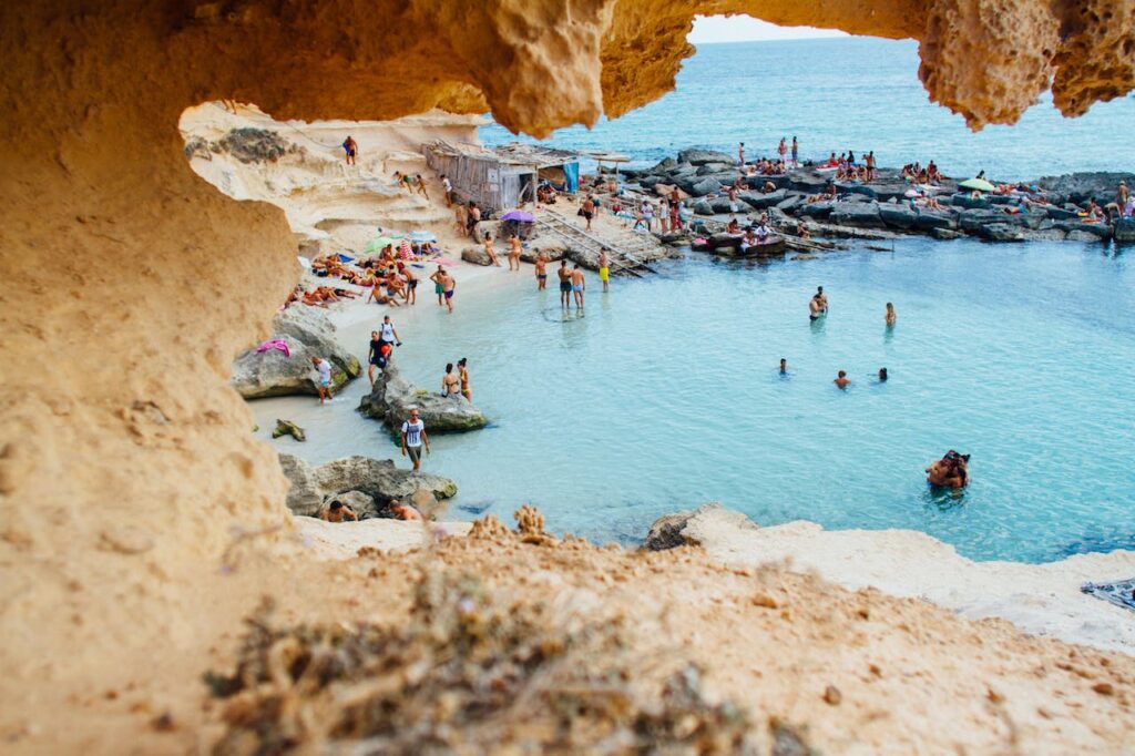 Beach scene with people swimming, lounging on rocks, and enjoying the sun by clear water and cliffs.