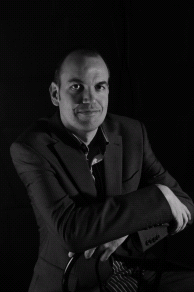 Professional in a suit seated with arms crossed, posed in a neutral, dark environment.