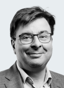 Professional man with glasses and a blazer, smiling against a neutral background.