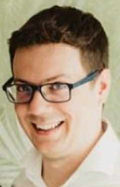 Smiling man with glasses, wearing a white shirt, in a neutral background setting.