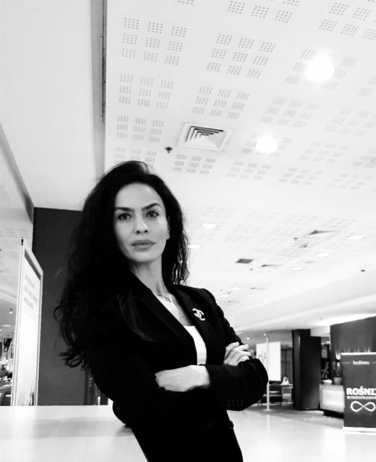 Legal professional standing confidently in a modern office space, arms crossed, wearing a black suit.