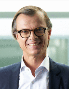 Smiling professional man wearing glasses in a business suit, with a blurred office background.