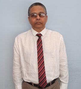 Legal professional in formal attire standing against a light wall, wearing glasses and a striped tie.