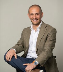 Smiling man in a suit sitting casually with arms crossed, against a neutral background.